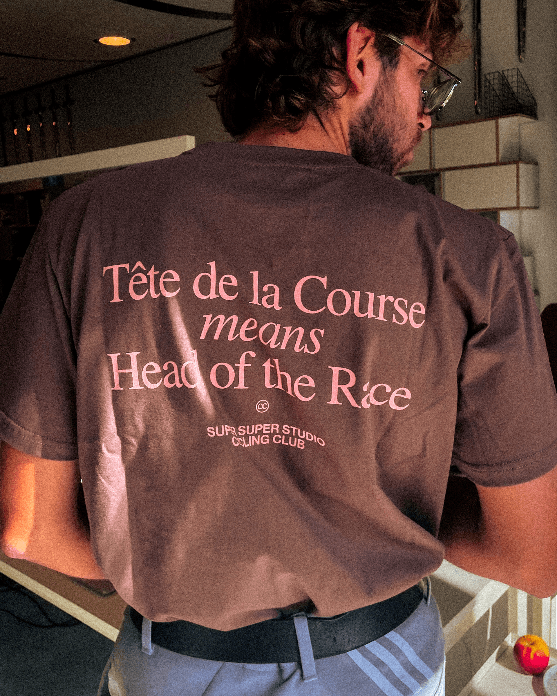 Back view of a brown T-shirt worn by a man, with pink text that reads 'Tête de la Course means Head of the Race' and 'Super Super Studio Cycling Club' at the bottom.